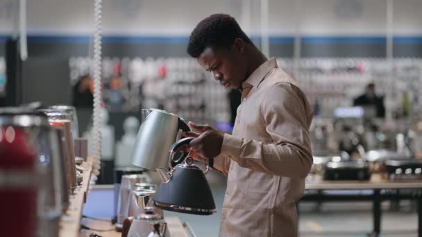 Jeune homme noir compare deux bouilloire électrique en magasin, tenant à la fois dans les mains et la pondération, visualisant différents modèles — Video