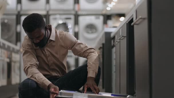 Молодой черный мужчина покупатель в маске для лица делает покупки в хозяйственном магазине во время пандемии, просматривая посудомоечную машину в торговой зоне — стоковое видео