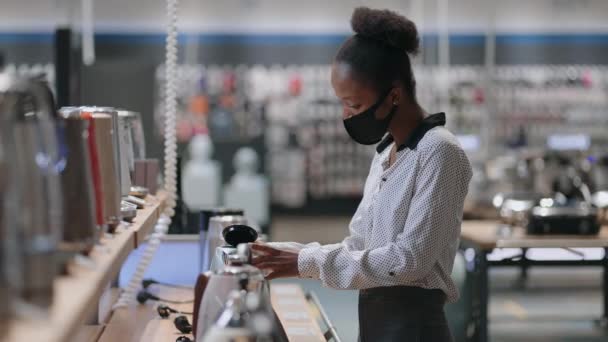 Yüz maskeli genç siyahi kadın ev aletleri süpermarketinde elektrikli çaydanlık ile mutfak aletini seçiyor. — Stok video