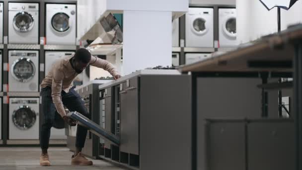 Negro chico está viendo lavavajillas en el área de comercio de la tienda de electrodomésticos modernos, abrir la puerta y examinar el interior — Vídeos de Stock