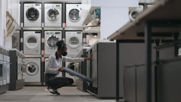 Giovane casalinga nera con maschera viso sta scegliendo lavastoviglie in elettrodomestici negozio, shopping nuove attrezzature per la cucina — Video Stock