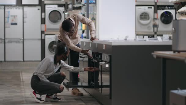 Família afro-americana está escolhendo forno em loja de ferragens no shopping, visualizando equipamentos dentro, comparando modelos — Vídeo de Stock