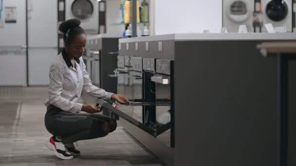 Jeune femme au foyer noire regarde four à l'intérieur avant d'acheter dans le magasin d'appareils ménagers, ouvrir la porte — Video