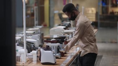 Afro-Amerikalı hırdavatçı hırdavatçıda, tek başına et öğütücüsünü seçiyor, mankenleri karşılaştırıyor, alışveriş merkezinin ziyaretçisi yüz maskesi takıyor.