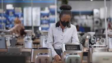 Genç siyahi ev hanımı ev aletleri dükkanında ekmek kızartma makinesine bakıyor. Afro-Amerikalı kadın, Coronavirüs 'ten korunmak için yüz maskesi takıyor.