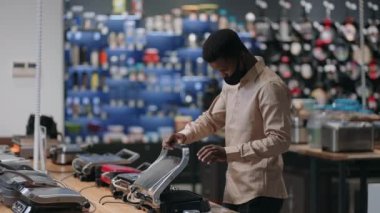 Yüz maskeli genç siyah adam ev aletleri mağazasından alışveriş yapıyor, elektrikli ızgara örneklerine bakıyor.