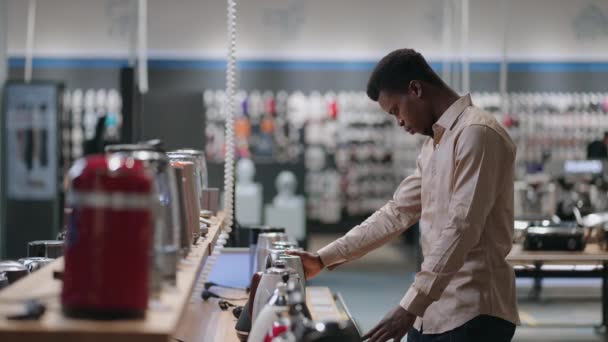 Hombre negro joven está eligiendo hervidor eléctrico, compras en la tienda de electrodomésticos en el centro comercial — Vídeos de Stock