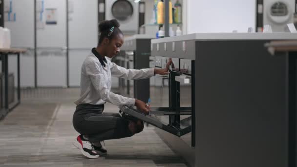 Jonge afro-Amerikaanse dame bekijkt moderne oven in hardware winkel, het kiezen van apparatuur voor nieuw appartement — Stockvideo