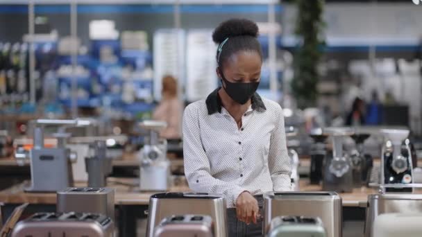 Jonge zwarte vrouw kiest broodrooster in huis apparaat winkel, shopper draagt gezichtsmasker voor de bescherming tegen Covid-19 — Stockvideo