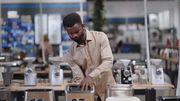Genç siyahi adam ev aletleri dükkanında tost makinesini seçiyor. Afro-Amerikalı adam hipermarkette covid-19 salgınında maske takıyor. — Stok video