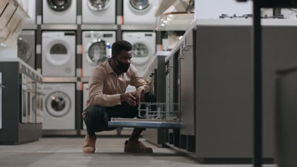 Hombre afro-americano con máscara facial está eligiendo lavavajillas en la tienda de electrodomésticos, abriendo la puerta y mirando dentro — Vídeos de Stock