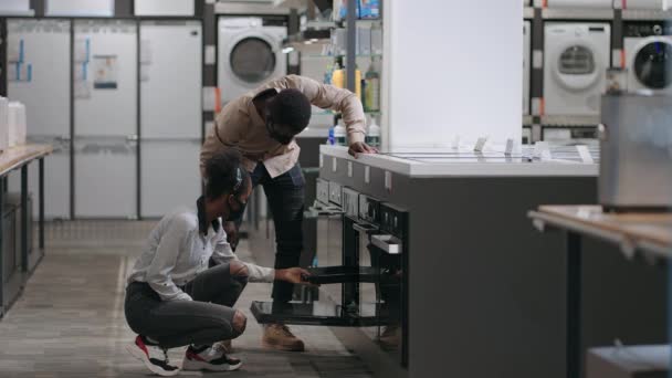 Молодые черные супруги делают покупки в магазине бытовой техники, выбирая духовку для домашней кухни — стоковое видео