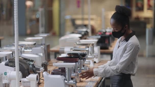 Winkelen in huishoudelijke apparaten winkel tijdens pandemie van het coronavirus, jonge zwarte vrouw is het kiezen van electro vleesmolen voor thuis keuken — Stockvideo