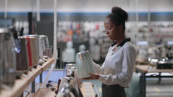 Afro-american lady is viewing electric kettle in home appliances store in mall, choosing model for home use — Stock Video