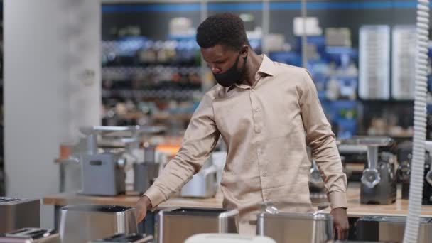 Jonge zwarte man met gezichtsmasker is het kiezen van broodrooster in hardware winkel, het bekijken van de tentoonstelling monster op plank in de handel gebied — Stockvideo