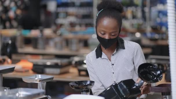 Joven mujer negra con máscara facial está de compras en la ferretería, la elección de electrodomésticos, la visualización de muestras de molinillo de carne eléctrica — Vídeo de stock