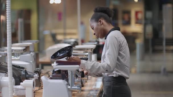 アフリカ系アメリカ人の女性が店内で電気肉グラインダーを見ている、若い主婦は台所用の家電製品を購入したい — ストック動画