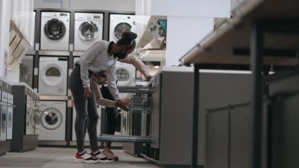 Чорний чоловік і жінка з масками для обличчя вибирають посудомийну машину в магазині побутової техніки, вивчають обладнання для кухні — стокове відео