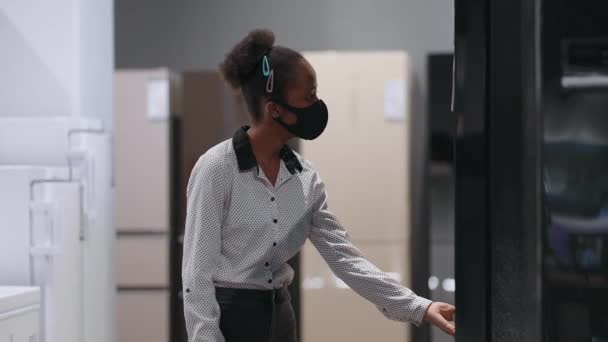 Ung svart kvinna med ansiktsmask väljer kylskåp med frys i hemmet apparat butik, visning utställning prov — Stockvideo