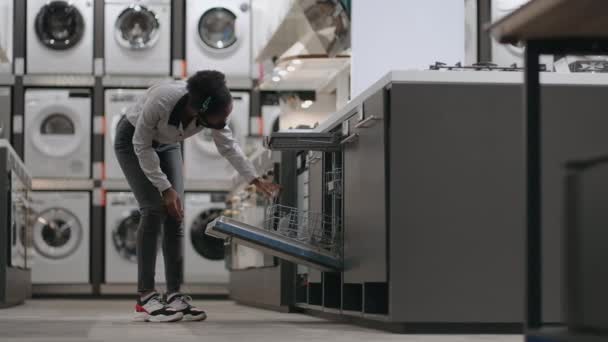 Покупки в магазине бытовой техники во время пандемии, молодая черная женщина в маске для лица смотрит посудомоечную машину — стоковое видео