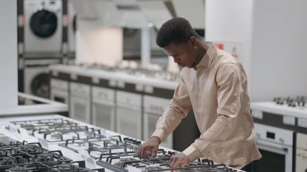 Mladý afro-americký chlap si vybírá sporák v obchodě s domácími spotřebiči, prohlížení povrchu vaření, svobodný muž je nakupování — Stock video