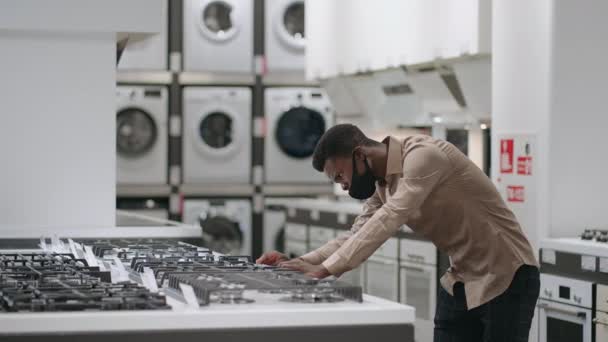 Jeune homme noir examine les caractéristiques du poêle dans le magasin d'appareils ménagers, lire les étiquettes et les étiquettes, homme seul avec masque facial — Video