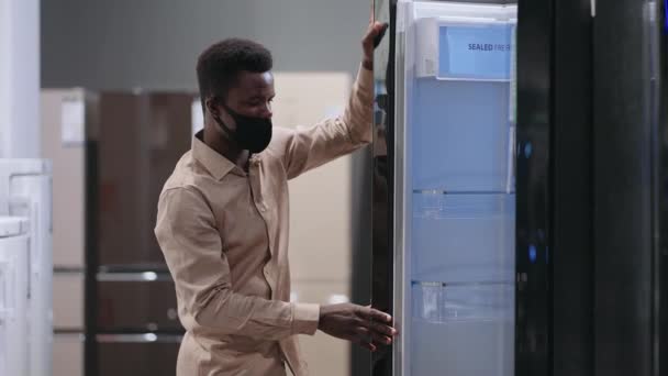 Erkek alışverişçi, ev aletleri dükkanındaki buzdolabına bakıyor. Mutfak için yeni ekipmanlar seçiyor. Afro-Amerikalı bir adamın orta boy portresi. — Stok video