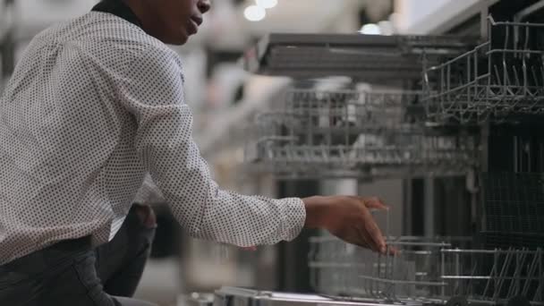 Joven mujer negra está eligiendo lavavajillas para la nueva cocina en el apartamento, compras en la tienda de electrodomésticos — Vídeo de stock