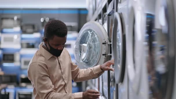 Hombre afroamericano con máscara facial está comprando lavadora en tienda de electrodomésticos durante la pandemia de covid-19, chico joven está viendo muestra de exposición — Vídeo de stock