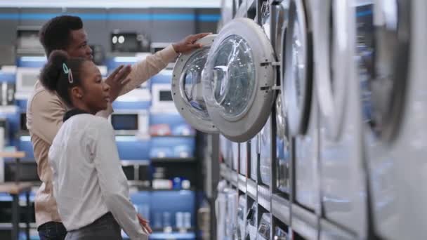 Cónyuges están eligiendo lavadora en la tienda de electrodomésticos, joven afroamericano hombre y mujer están viendo muestra de exposición — Vídeo de stock