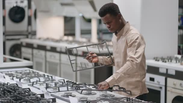 Ernster junger Mann kauft Gasherd im Geschäft für Haushaltsgeräte, untersucht Kochfläche, afroamerikanischer Mann — Stockvideo