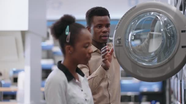Sezdaný pár nakupuje v obchodě s domácími spotřebiči, afro-američan muž a žena si prohlíží exponát ukázka pračky — Stock video