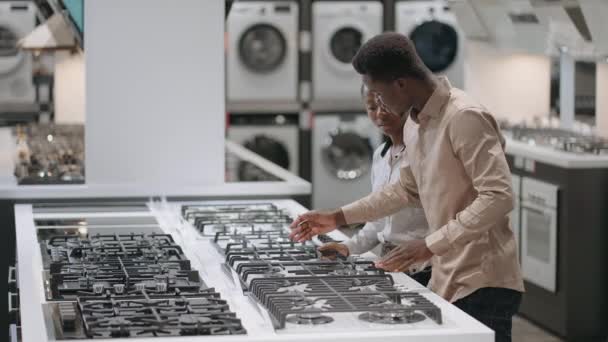 Spouses are discussing buying of new equipment for kitchen, afro-american pair is shopping in department with gas cookers in home appliance stores in mall — Stock Video