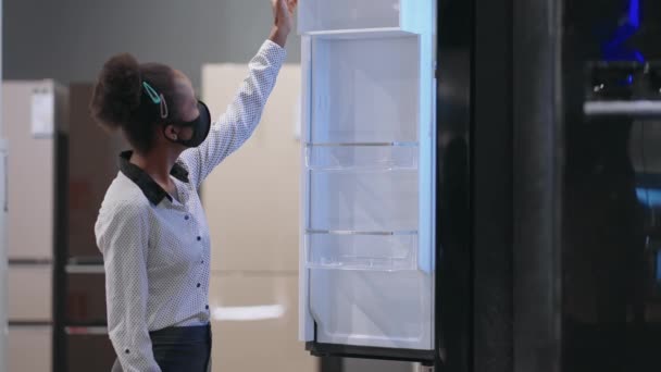 Jeune femme au foyer noire avec masque facial choisit réfrigérateur dans le magasin d'appareils ménagers, visualisation de l'échantillon d'exposition — Video