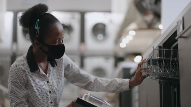 Jeune afro-américaine dame avec masque facial achète lave-vaisselle dans le magasin d'appareils ménagers, visualisation examen échantillon d'exposition — Video