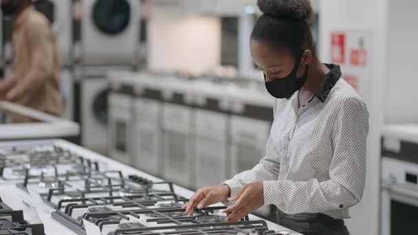 Tiendas de electrodomésticos en el centro comercial, la gente está de compras en el departamento con estufas, señora afroamericana con máscara facial está viendo la superficie de cocción — Vídeos de Stock