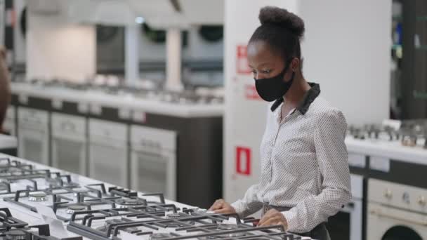 Jonge vrouwelijke shopper met gezichtsmasker is het kiezen van nieuwe gasfornuis in huishoudelijke apparaten winkel, onderzoek kookoppervlak, afro-Amerikaanse huisvrouw — Stockvideo