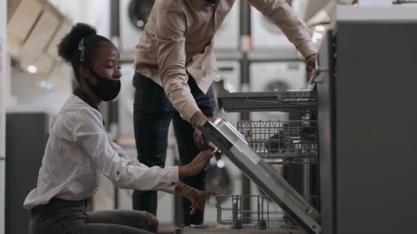 Jóvenes afroamericanos cónyuges están eligiendo lavavajillas en la tienda de electrodomésticos, hombre y mujer están viendo el equipo para la cocina — Vídeos de Stock