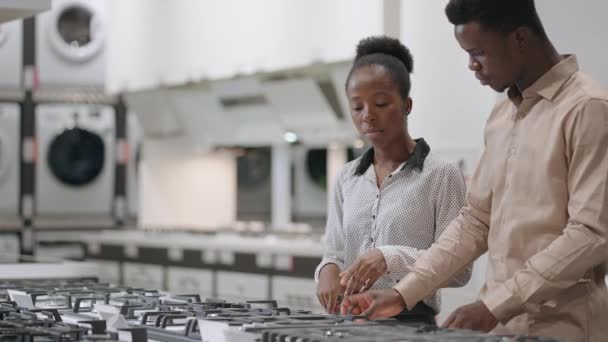 Jonge echtgenoten kiezen gasfornuis in huishoudelijke apparaten winkels in winkelcentrum, zwarte man en vrouw zijn het bekijken van het kookoppervlak en bespreken — Stockvideo