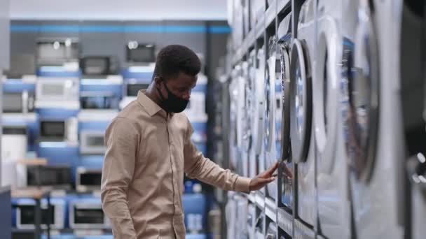 Молодий чорний чоловік переглядає виставкові зразки пральних машин у торговому залі магазину побутової техніки під час пандемії — стокове відео