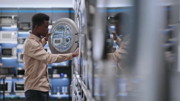 Visitante masculino del centro comercial está eligiendo la lavadora en la tienda de electrodomésticos, chico afroamericano joven está viendo muestra de exposición en la sala de comercio — Vídeo de stock
