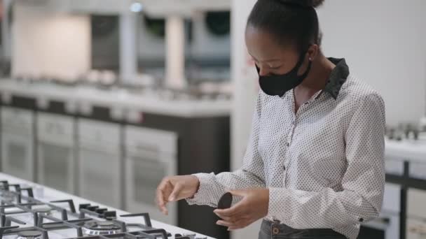 Jeune femme noire avec masque facial choisit cuisinière à gaz dans les magasins d'appareils ménagers dans le centre commercial, magasinage seul à la période pandémique, regarder des échantillons d'exposition — Video