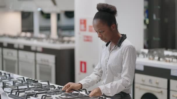 Compras en tiendas de electrodomésticos en el centro comercial, mujer afroamericana joven está viendo muestras de exposición de estufa, la elección de nuevos equipos para la cocina — Vídeos de Stock