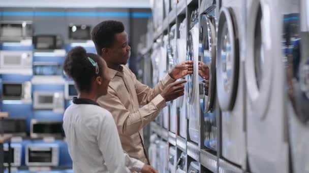 Відвідувачі торгового центру вибирають пральну машину в магазині побутової техніки, молоді афроамериканські подружжя переглядають виставковий зразок у торговому залі — стокове відео