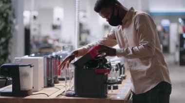 Afro-Amerikalı adam ev aletleri dükkanından otomatik kahve makinesini seçiyor. Maskeli genç adam alışveriş yapıyor.