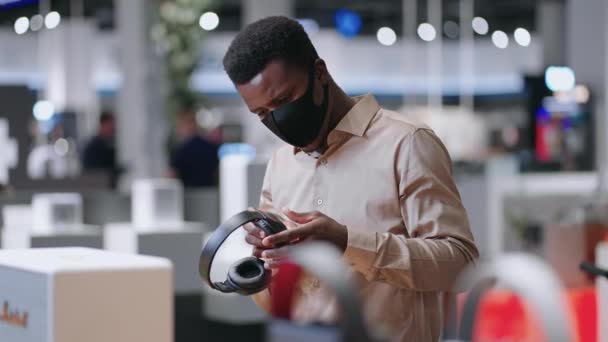 Młody czarny facet ogląda nowy model słuchawek bezprzewodowych w hali handlowej sklepu ze sprzętem audio, średni portret kupującego — Wideo stockowe