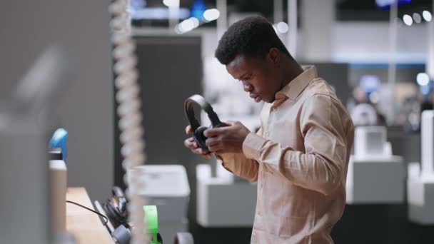 Afro-americano cara está testando fones de ouvido modernos na loja de equipamentos de áudio, retrato médio de homem comprador na sala de negociação — Vídeo de Stock