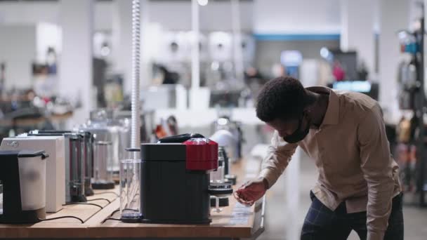 Jonge afro-Amerikaanse man kiest koffiezetapparaat in huis apparaat winkel, jonge man met gezichtsmasker onderzoekt tentoonstelling monster in hardware winkel — Stockvideo