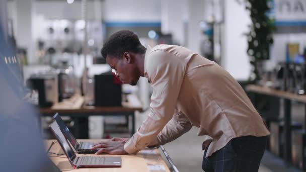 Homem afro-americano está escolhendo laptop na loja de eletrônicos, visualização de amostras de exposição no salão de negociação — Vídeo de Stock