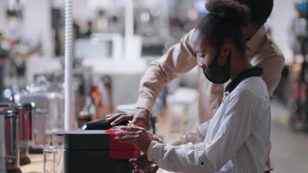 Афро-американська пара з масками для обличчя вибирає автоматичну кавоварку в магазині побутової техніки, переглядаючи виставкові зразки — стокове відео